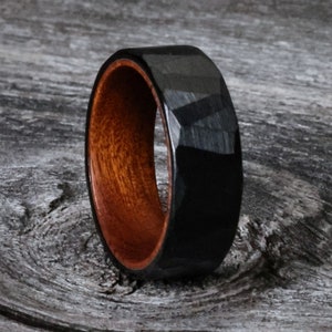 May include: A black faceted ring with a dark wood interior. The ring has a geometric design and a polished finish. The wood interior has a reddish-brown color. The ring is likely made of wood and metal.