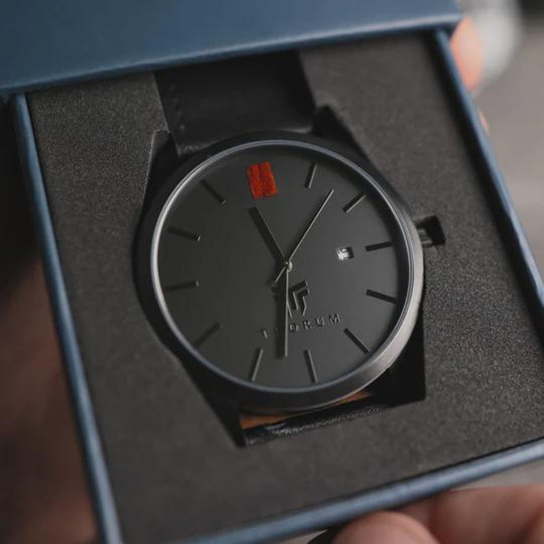 May include: A black wristwatch with a black leather band is presented in a blue box. The watch face is dark gray with black hands and markers, a small date window, and a red accent. The brand name "TORUM" is printed on the watch face.