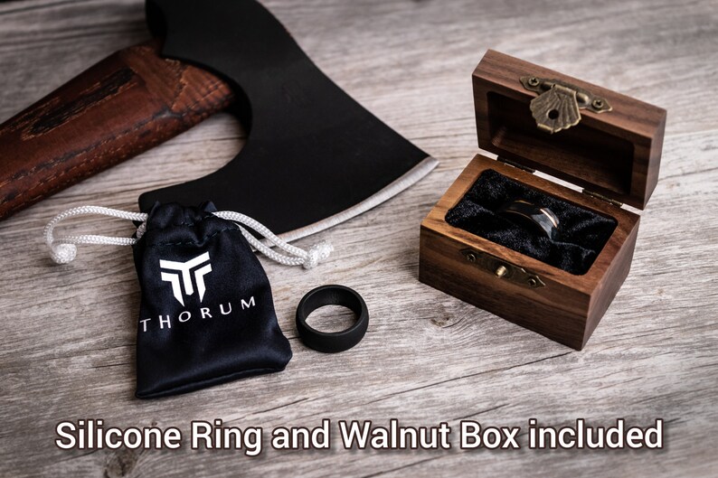 May include: A black axe head with a wooden handle, a black silicone ring, a black drawstring bag with a white logo, and a wooden box with a ring inside. The text "Silicone Ring and Walnut Box included" is at the bottom.