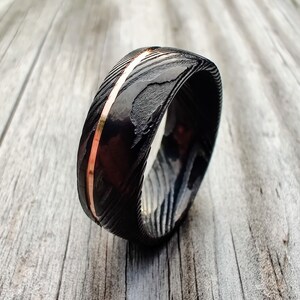 May include: A black and copper-colored ring with a textured surface and a copper-colored stripe down the center. The ring is set against a light wood grain background.