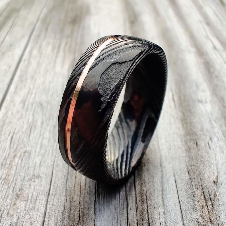 May include: A black and copper-colored ring with a textured surface and a copper-colored stripe down the center. The ring is set against a light wood grain background.