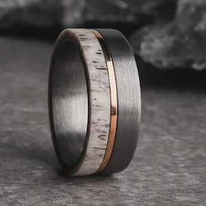 May include: A close-up of a ring with a dark gray band, a white and brown center inlay, and a thin gold-colored stripe. The ring is made of metal and has a brushed finish. The ring is a piece of jewelry.