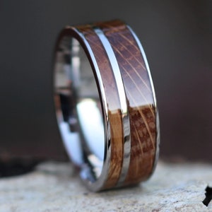 Puede incluir: Primer plano de un anillo con una banda plateada pulida y una incrustaci&oacute;n central de madera marr&oacute;n claro. Una fina franja plateada recorre el centro de la incrustaci&oacute;n de madera. El anillo tiene una superficie brillante y reflectante.