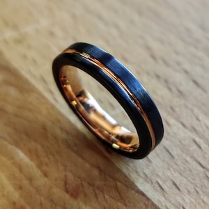 May include: A close-up of a black ring with a thin, rose gold-colored stripe down the center. The inside of the ring is also rose gold. The ring is on a wooden surface.