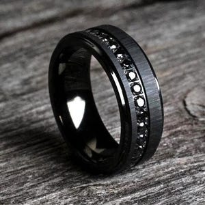 May include: Black ring with a brushed metal band and a row of black gemstones. The ring has a polished inner band and is set against a wooden surface.