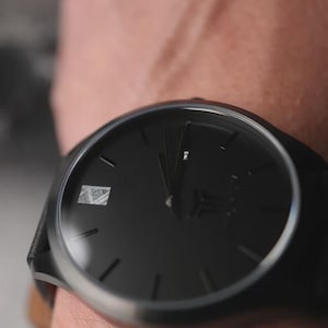 May include: A black wristwatch with a dark face and silver bezel. The watch has a minimalist design with simple hour markers and hands. The watch band appears to be black leather.