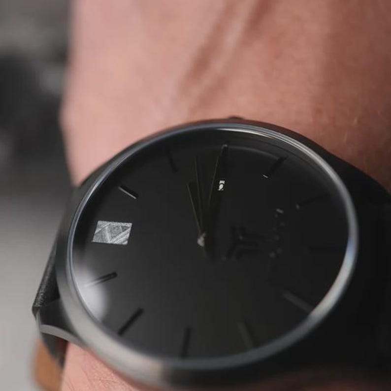 May include: A black wristwatch with a dark face and silver bezel. The watch has a minimalist design with simple hour markers and hands. The watch band appears to be black leather.