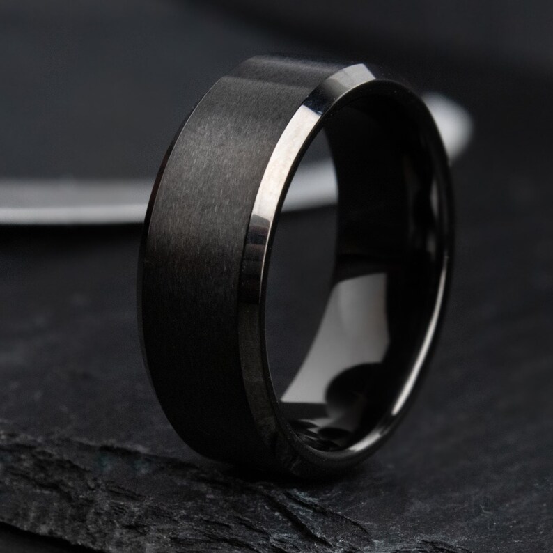 May include: A black ring with a brushed metal band and a polished interior. The ring features a silver-coloured edge and a modern design. The ring is displayed on a dark, textured surface. The ring is a piece of jewellery.