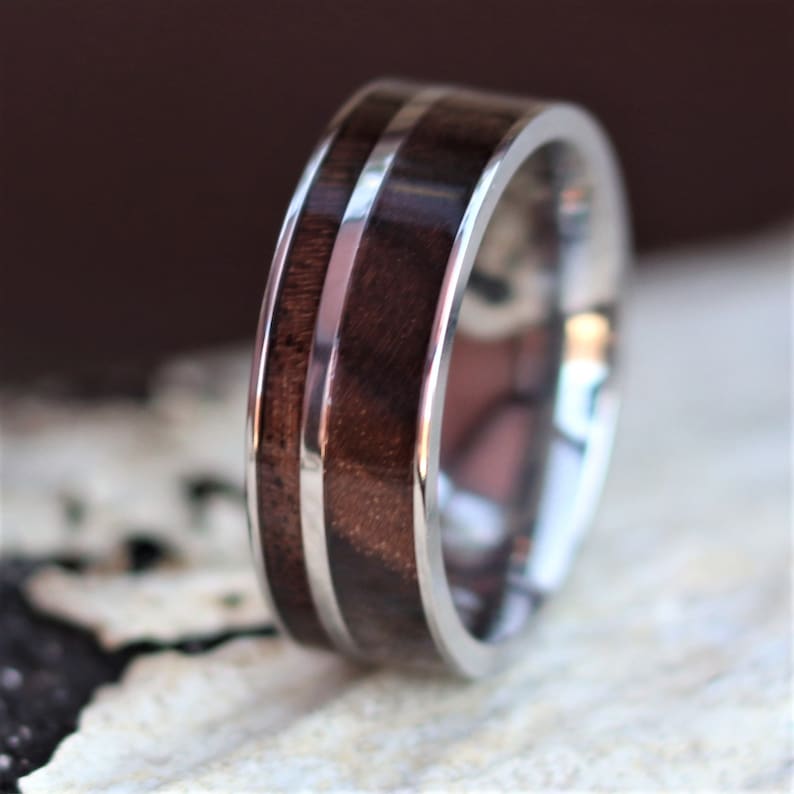 May include: A close-up of a silver ring with a dark brown wood inlay. The ring has a polished finish and a band of silver running through the center of the wood. The ring is a classic design.
