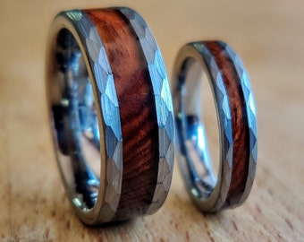 May include: Two wedding rings with a hammered silver band and a dark wood inlay. The rings are set on a wooden surface. The wood grain is visible in the inlay. The rings are in focus.