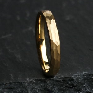 May include: A gold-colored ring with a hammered texture. The ring is set against a dark background, highlighting its metallic sheen. The ring's surface has a textured, irregular pattern.