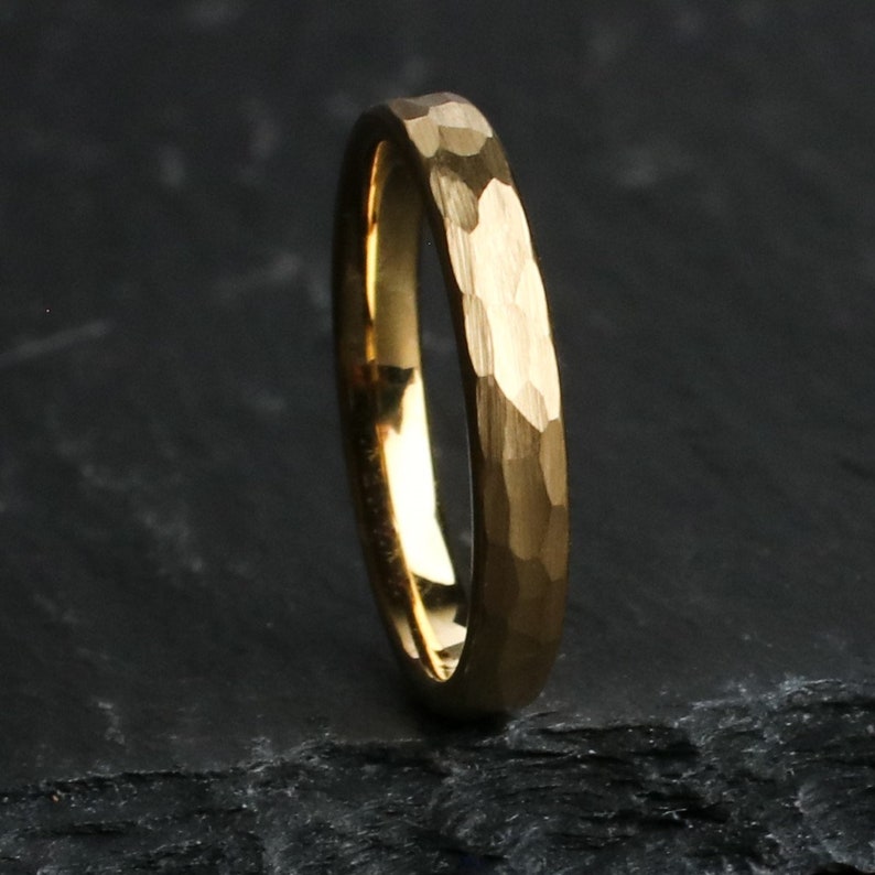 May include: A gold-colored ring with a hammered texture. The ring is set against a dark background, highlighting its metallic sheen. The ring's surface has a textured, irregular pattern.
