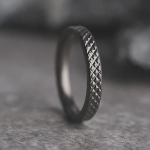 May include: A close-up of a dark gray ring with a textured, diamond-patterned surface. The ring is made of metal and has a thin band. The background is blurred, highlighting the ring's intricate design.