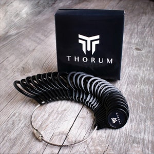 Puede incluir: Juego de medidores de anillos negros con un alambre plateado y una caja negra con la palabra "THORUM" en blanco. El juego de medidores de anillos está sobre una superficie de madera.