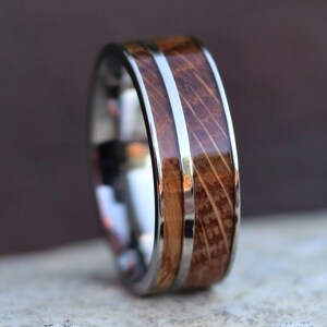 Puede incluir: Primer plano de un anillo con dise&ntilde;o de barril de madera. El anillo tiene una banda plateada con una incrustaci&oacute;n de madera. La madera tiene un aspecto marr&oacute;n y texturizado. El anillo probablemente est&aacute; hecho de acero inoxidable y madera.