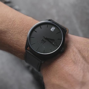 May include: A black wristwatch with a leather band. The watch face is black with minimalist hour markers and hands. The brand name "THORUM" is printed at the bottom of the watch face. The watch is on a wrist.