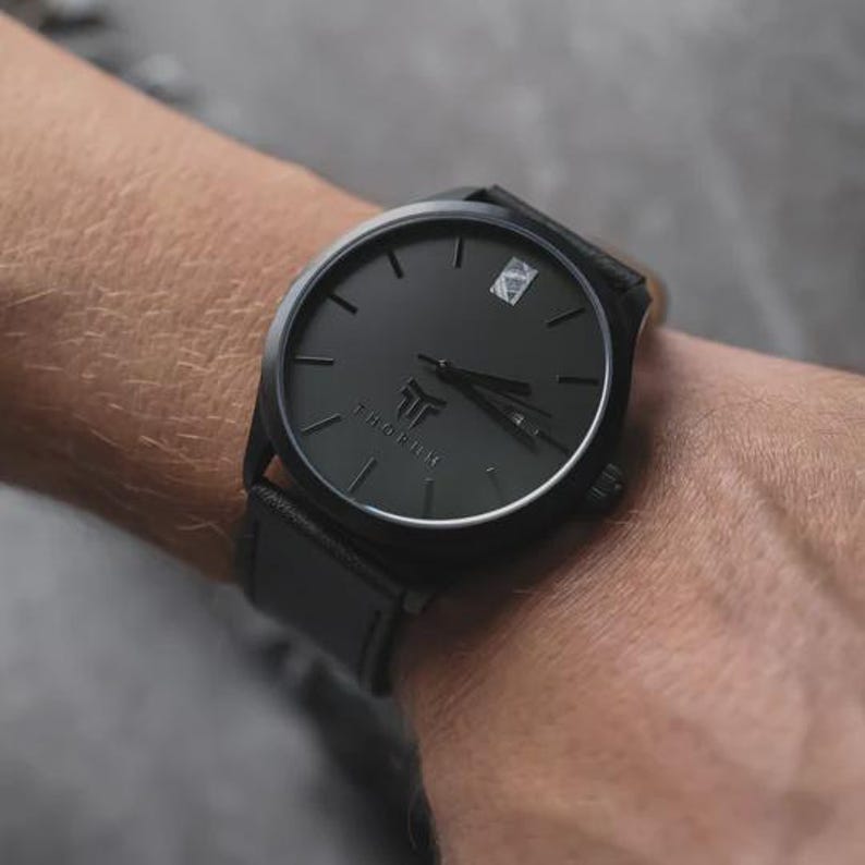 May include: A black wristwatch with a leather band. The watch face is black with minimalist hour markers and hands. The brand name "THORUM" is printed at the bottom of the watch face. The watch is on a wrist.