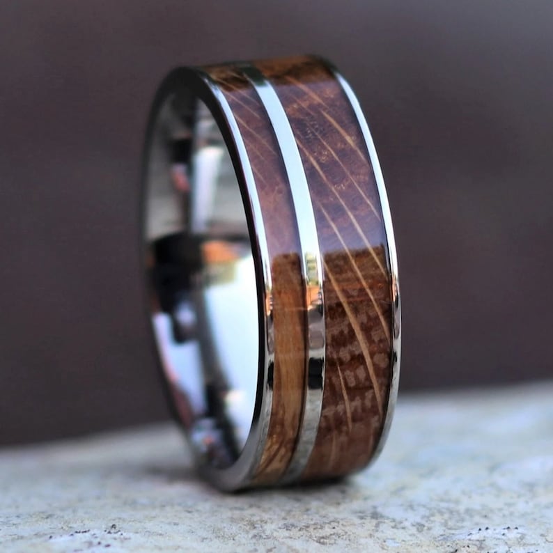 Puede incluir: Primer plano de un anillo con una banda plateada pulida y una incrustaci&oacute;n de madera. La madera presenta un patr&oacute;n de vetas natural y est&aacute; enmarcada por una fina franja plateada. El anillo se encuentra sobre un fondo borroso.