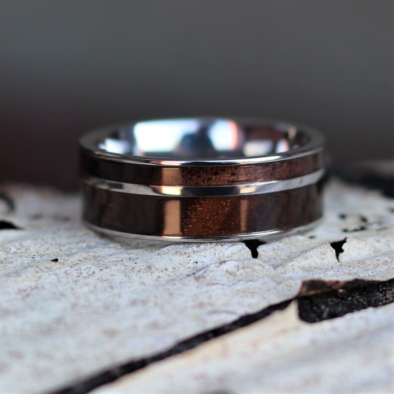 May include: A close-up of a ring with a dark wood inlay and a silver-colored metal band. The wood has a rich, brown color, and the metal has a polished, reflective surface. The ring is a band style, with a simple, elegant design.
