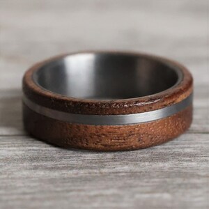 May include: A close-up of a wooden ring with a silver band and a dark metal interior. The ring has a natural wood grain texture and a polished finish. The ring is set against a light gray wooden surface.