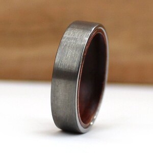 May include: A close-up of a ring with a brushed grey metal exterior and a dark reddish-brown wooden interior. The ring has a smooth, rounded profile, showcasing the contrast between the metal and wood.