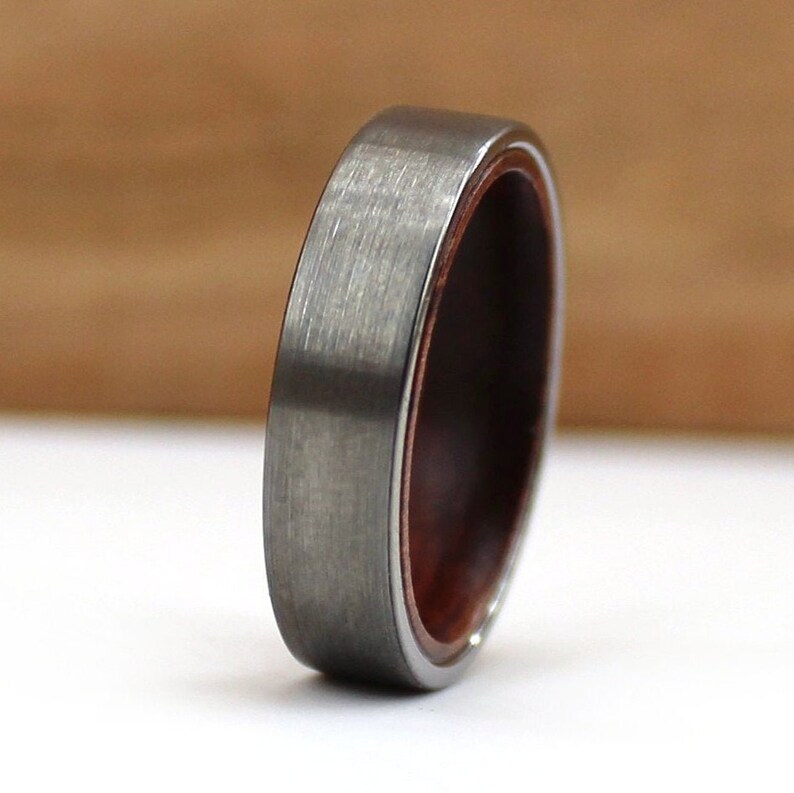 May include: A close-up of a ring with a brushed grey metal exterior and a dark reddish-brown wooden interior. The ring has a smooth, rounded profile, showcasing the contrast between the metal and wood.