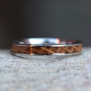 May include: A silver ring with a wooden inlay. The ring features a polished silver band with a brown wooden center. The wood has a diagonal grain pattern and small, light-colored spots.