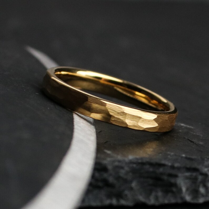 May include: A gold-colored ring with a hammered texture. The ring is set against a dark background, with a subtle, curved silver line. The ring's surface reflects light, highlighting its textured design. The ring is a simple band, likely for everyday wear.