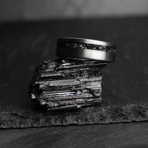 May include: A dark grey ring with a black stone inlay sits on a black, textured mineral. The ring has a brushed metal finish and a band of small, dark stones. The background is a dark, blurred surface, creating a dramatic contrast.