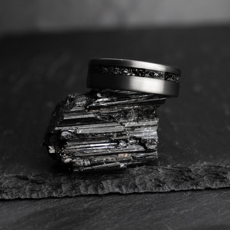 May include: A dark grey ring with a black stone inlay sits on a black, textured mineral. The ring has a brushed metal finish and a band of small, dark stones. The background is a dark, blurred surface, creating a dramatic contrast.