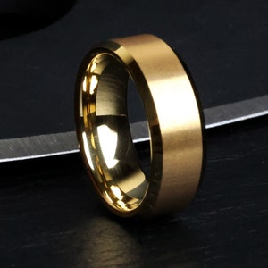 May include: A gold-colored ring with a flat top and beveled edges. The ring has a smooth, polished interior and a matte finish on the exterior. The ring is likely made of metal and is designed to be worn on a finger.
