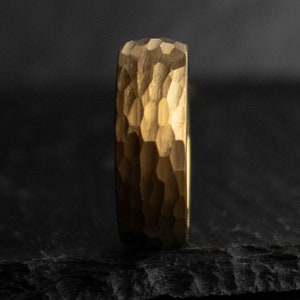 May include: A gold-colored ring with a textured, hammered surface. The ring's band has a geometric pattern, and the overall design is simple and elegant. The ring is shown against a dark background.