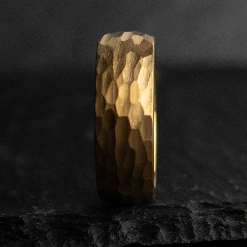 May include: A gold-colored ring with a textured, hammered surface. The ring's band has a geometric pattern, and the overall design is simple and elegant. The ring is shown against a dark background.