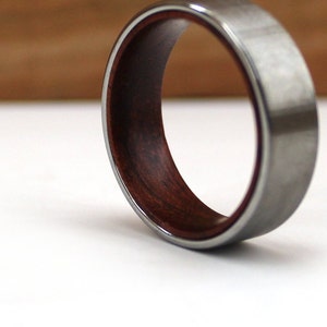 May include: A close-up shot of a ring with a silver-colored exterior and a dark reddish-brown wooden interior. The ring is set against a white background, with a blurred wooden surface in the background. The ring's design features a contrasting combination of materials.