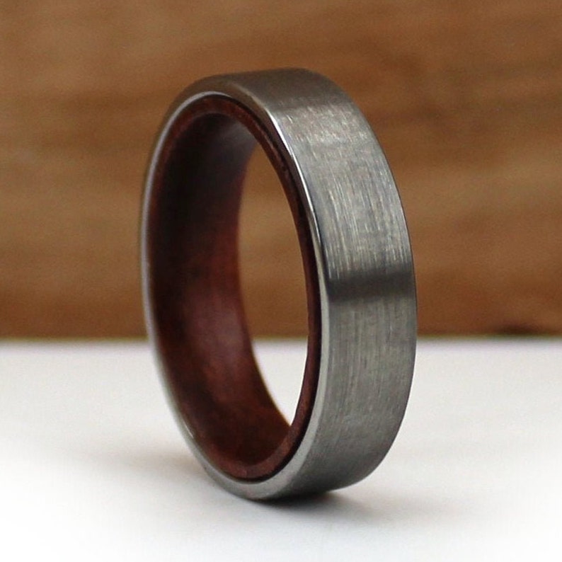 May include: A close-up of a brushed metal ring with a dark wood interior. The ring has a flat profile and a silver-grey exterior. The wood interior is a deep reddish-brown colour, creating a contrast with the metal.