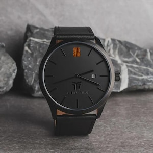 May include: A black wristwatch with a leather band. The watch face is black with minimalist hour markers and hands. The brand name "THORUM" is printed at the bottom of the watch face. The watch has a small date window and a textured crown.