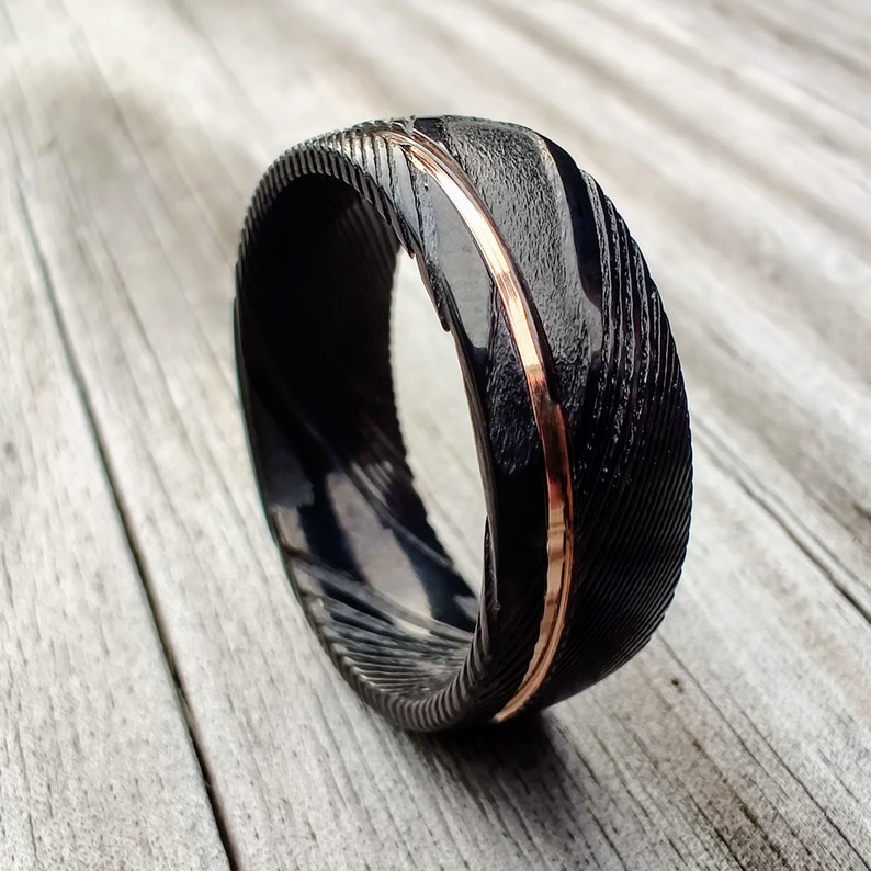 May include: A close-up of a black ring with a textured surface and a thin, rose gold band. The ring is set against a light wood grain background, highlighting the contrast between the dark metal and the warm gold accent.