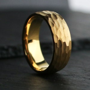 May include: A gold-colored ring with a textured, hammered surface. The ring has a smooth, polished interior and a band width of approximately 6 mm. The ring is set against a dark background, highlighting its metallic sheen.