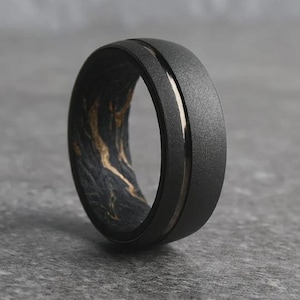 May include: A black ring with a textured matte finish and a thin, silver-colored inlay. The interior of the ring features a marbled pattern in shades of black and gold. The ring is a close-up shot against a gray background.