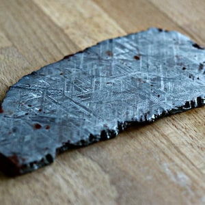May include: A slice of a meteorite with a distinctive geometric pattern. The dark grey stone has a rough, uneven edge and is displayed on a wooden surface. Small reddish-brown spots are visible on the surface.