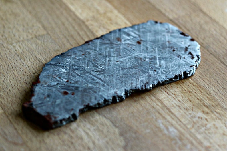 May include: A slice of a meteorite with a distinctive geometric pattern. The dark grey stone has a rough, uneven edge and is displayed on a wooden surface. Small reddish-brown spots are visible on the surface.