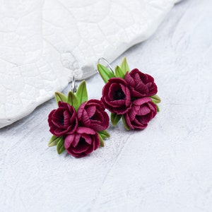 May include: A pair of burgundy flower earrings with green leaves. The earrings are made of polymer clay and have a silver hook.