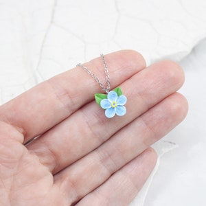 May include: A delicate silver chain necklace with a blue forget-me-not flower charm. The flower has five petals and green leaves.