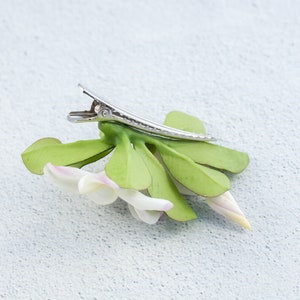 May include: A white flower with green leaves attached to a silver hair clip. The flower has a pink center and is made of fabric.