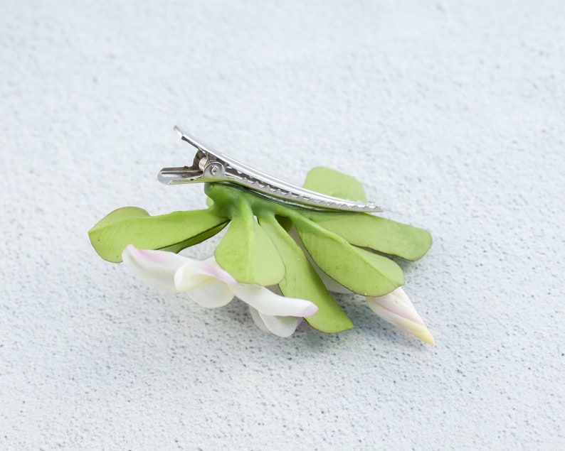 May include: A white flower with green leaves attached to a silver hair clip. The flower has a pink center and is made of fabric.