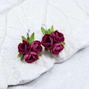 May include: A pair of earrings featuring three burgundy flowers with green leaves. The earrings are made of polymer clay and have silver hooks.