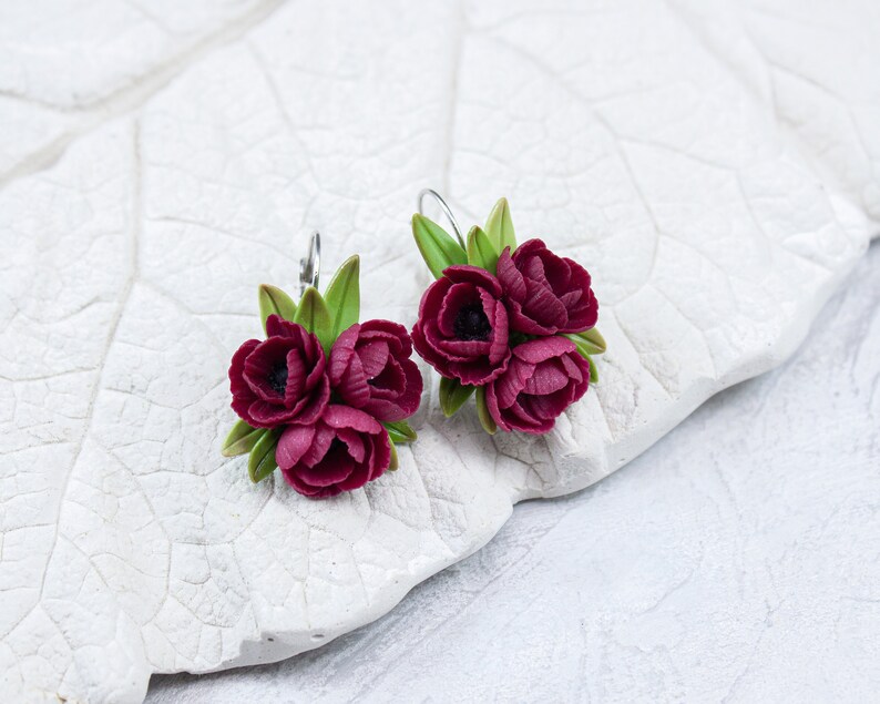 May include: A pair of earrings featuring three burgundy flowers with green leaves. The earrings are made of polymer clay and have silver hooks.