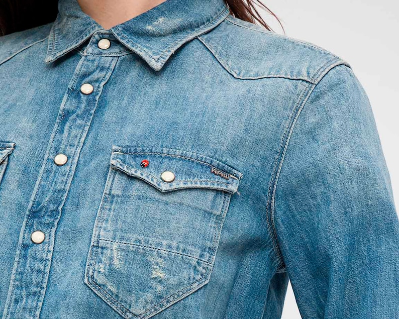 May include: A light blue denim shirt featuring a classic collar, snap button closures, and two chest pockets. The left pocket displays a small red ladybug pin and a metal tag with the text RAW. The fabric has a distressed, faded wash finish.