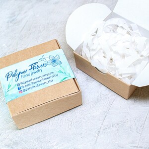 May include: A small, brown cardboard box with a white and blue label that says "Polymer Flowers Floral Jewelry" and includes contact information for the shop. The box is filled with white tissue paper.