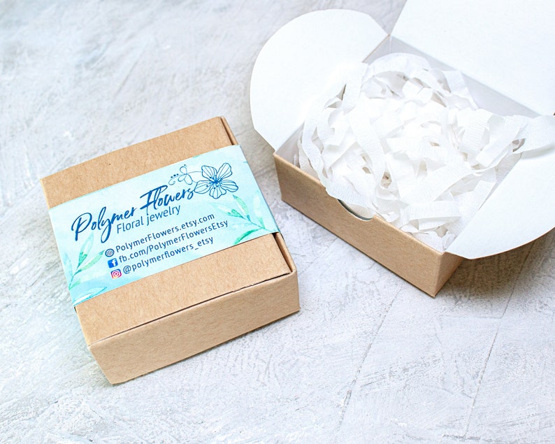 May include: A small, brown cardboard box with a white and blue label that says "Polymer Flowers Floral Jewelry" and includes contact information for the shop. The box is filled with white tissue paper.